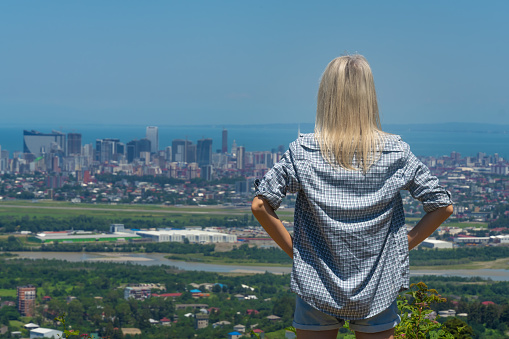 Young blonde woman in a plaid shirt looks at the city from the height of the mountain from the back. Girl with blonde hair looks down at the city, the sea. Concept of travel and wanderlust.
