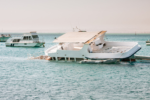 A white boat resting in a harbor, surrounded by other boats in the background. The boat appears to have suffered damage and is partially submerged in the water. It has a flat roof and a cabin area that has collapsed partially. The boat leans to the left, and its front is submerged in the light blue water. The sky above is a hazy shade of blue, adding to the overall scene.