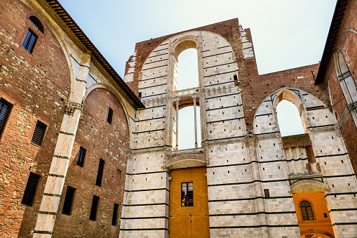 The unfinished structure of the new cathedral on the right side of the Duomo in the medieval heart of Siena, in Tuscany. The construction of the Metropolitan Cathedral or Duomo dedicated to the Assumption of Mary was completed between 1215 and 1348, becoming one of the religious architectural masterpieces of the Romanesque and Italian Gothic style. But in 1339 it was decided to expand the Cathedral, leaving the existing structure as a transept of the new construction. Unfortunately the new and larger cathedral was not completed and the structure of the unfinished main nave (in the photo) is today called the Duomo Nuovo. Siena is one of the most beautiful Italian cities of art, in the heart of the Tuscan hills, famous for its immense artistic and historical heritage and for the Palio, where the seventeen historical districts of the city compete every year in Piazza del Campo in one of the oldest horse racing in the world. Since 1995 the historic center of Siena has been declared a World Heritage Site by UNESCO. Wide angle mage in high definition quality.