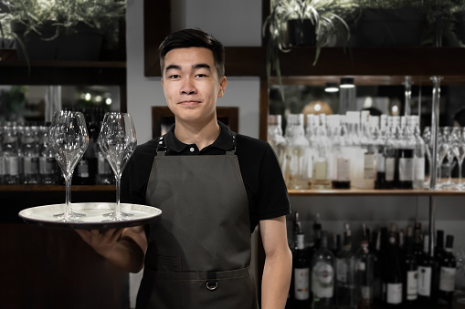 Close-up portrait of a smiling Asian waiter dressed in a monochromatic uniform holing a rounded tray with wineglasses. Young server guy wearing dark t-shirt and green apron with straps standing at bar counter and carrying salver with glassware. Blurred background. Coffee shop concept.