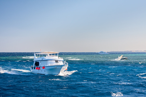 A white yacht sailing on a blue ocean. The yacht is seen moving towards the right side of the image, leaving a trail of white foam behind it. It features a red lifebuoy on the side. In the background, another yacht can be observed, also in motion, heading towards the left side of the image. The ocean appears choppy with small waves, while the sky above is clear and blue. The horizon is visible in the background.