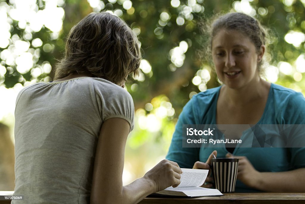 Friends, Coffee, and Discussing a Book Two young women studying the Bible or some other book and talking about it outside on a warm summer day.  (Focus is on woman's hand turning the page) Bible Stock Photo