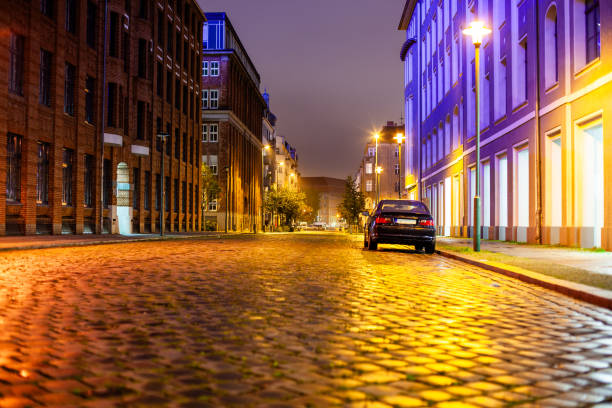 A cobblestone street at night in a European city. The street is lined with brick buildings on both sides, with a black car parked on the right side of the street. The street is lit by street lamps, which create a warm glow on the cobblestones. The sky is dark and cloudy, and the buildings in the background fade into the distance.