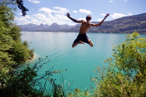 Young man leaps from a cliff into a blue Alpine lake below on a hot summer day. 