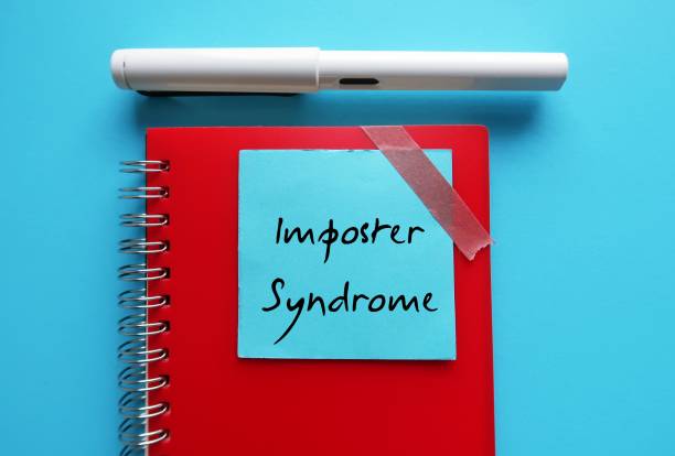 istock Blue note on red notebook with handwritten text IMPOSTER SYNDROME, feeling that one less capable than the people around with fear of failure and other people will find out a fake 1573763444