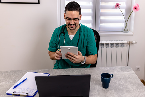 Cheerful smiling mid adult Caucasian male nurse in uniform sitting in front of laptop using tablet