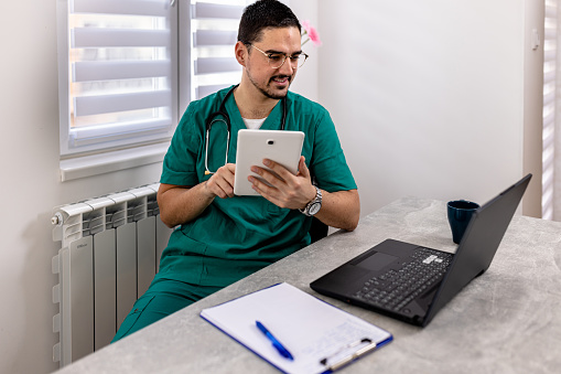Focused cheerful mid adult Caucasian male doctor sitting in front of laptop holding tablet finishing some work