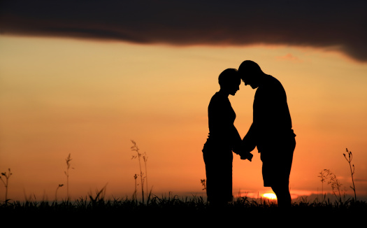 A couple sharing a moment at sunset. Themes include marriage, romance, love, relationships, couple, heterosexual, sex, intimacy, anniversary, honeymoon, sharing, talking, bonding, holding, hands, valentine's day, side view, unrecognizable people, two people, rural, meadow, outside, and embracing. 