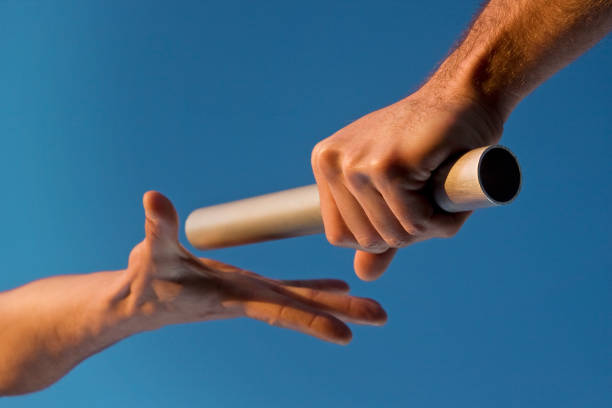 Two Hands Passing Relay Baton Race Low angle close-up of two hands passing a relay baton, with focus on the hand holding the baton, against a clear blue sky. Warm tones highlight the fact that the image was shot at sunset. Horizontal format. relay stock pictures, royalty-free photos & images