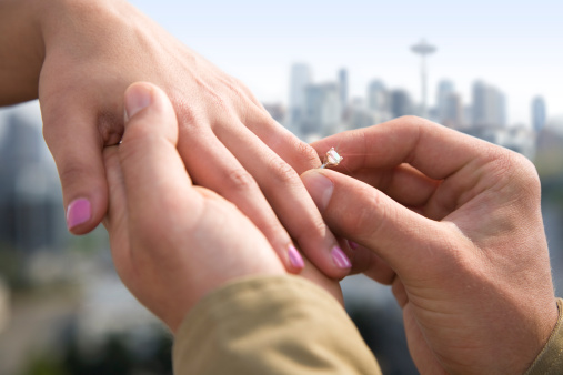 A man proposed as he slips a diamond ring onto the finger of a woman with the city of Seattle, Washington in the background.