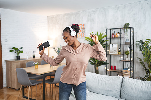 Black woman having fun listening to music on her wireless headphones and dancing in the living room