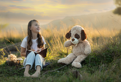 Young, smiling girl sitting in nature at sunset with her teddy writing in her book