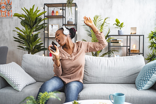 Black woman having fun listening to music on her wireless headphones and singing in the living room