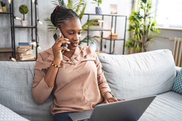 istock Black woman working on her laptop and talking on a phone call while sitting on the sofa in her living room 1573803692