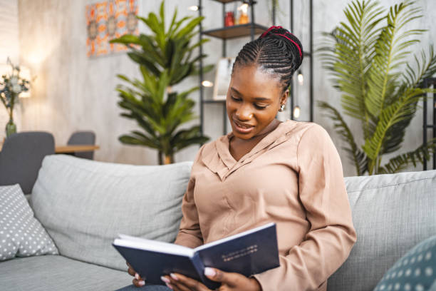 istock Black woman sitting on her living room sofa reading a book 1573804073