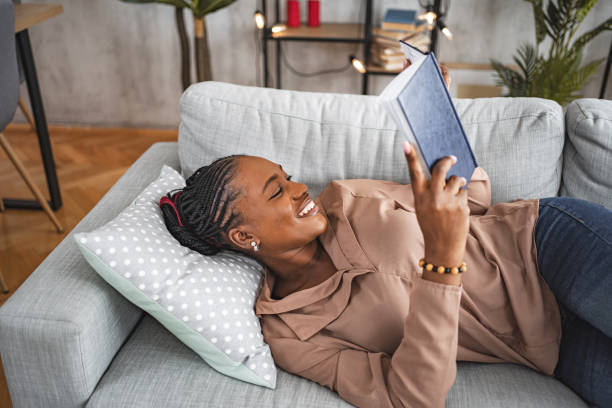 istock Black woman reading a book while lying on her living room sofa 1573804260