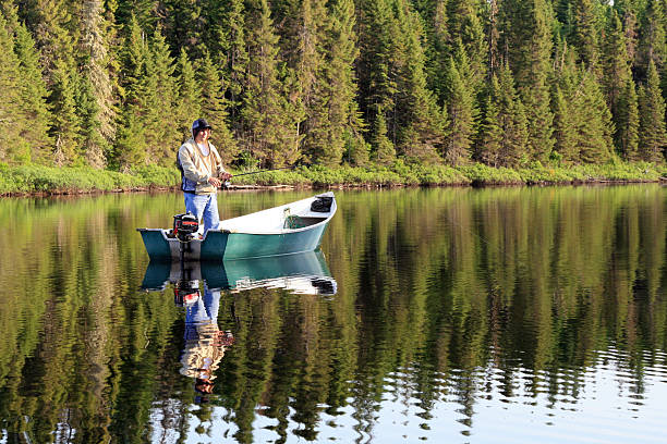 DSLR picture of a fisherman fishing on a quiet lake. The fisherman is standing up in a motor boat and catching a fish. His reflection is visible on the lake. He is surrounded by a forest of coniferous.  