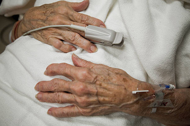 senior woman weathered old wrinkled hands with IV drip on wrist and medical equipment oxygen sensor (pulse oximeter) on finger, is a patient in ER emergency room hospital bed
