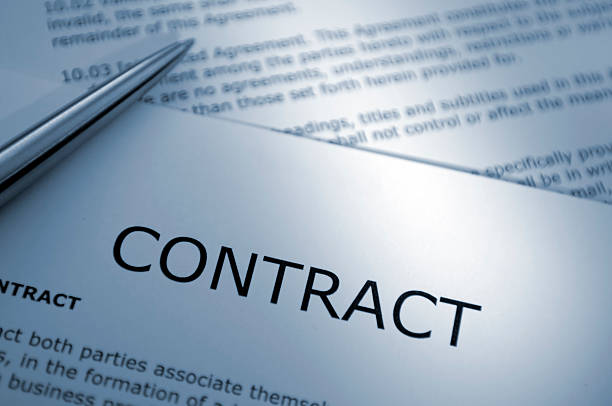 A macro close up blue toned image of a ballpoint pen resting on sheets of legal contract paperwork documents. Show with  shallow depth of field with focus on the word ‘Contract’ document title. All contact text is generic and fake.   