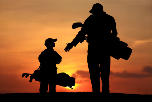 A father gives his son a high five on the golf course. Silhouette. Additional themes of the image are father's day, relationship, bonding, care, togetherness, love, single parent, father, parenting, child, boys, fun, sport, game, playing, leisure, recreation, walking, encouragement, role model, aspirations, dads, and congratulating. Rear view. Unrecognizable people. Boy is elementary age. 