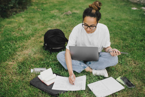 istock Young woman student studying using her books, laptop and notebook, sitting on grass. 1573866943