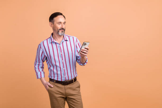 istock Portrait of positive handsome elderly man hold use smart phone empty space offer promo isolated on beige color background 1573868921