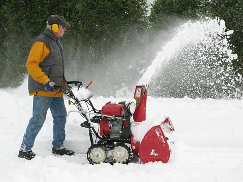 A man in his 50s using a snow blower with caterpillar tracks to clear the snow from his driveway after a heavy fall.