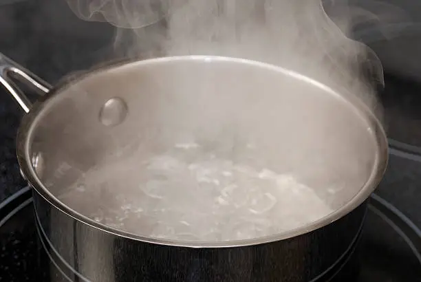 Water bubbles and boils in a pot on the stove.  Water looks soft due to the steam.  Pot is stainless steel on a black stove top.  Lots of steam.