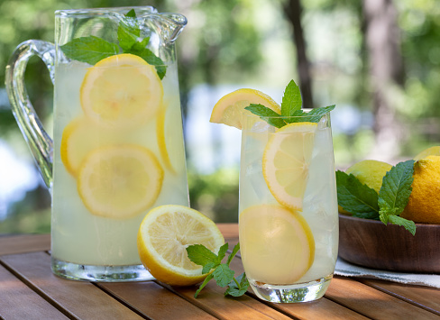 Lemonade in glass and pitcher with mint and sliced lemons outdoors on wooden patio table and nature background