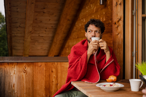A shot of a male sitting at a table on the balcony of a wooden chalet in the Alpine town of Garmisch, Bavaria. He is holding a cup of coffee, looking off camera into the distance, wrapped in a blanket. On the table is a bowl of cereal, tangerine and his partner's cup of coffee, who sits opposite off-camera.