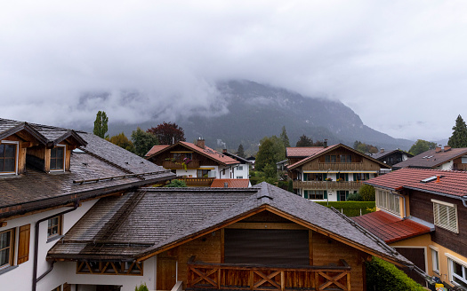 A wide shot looking across chalet rooftops from a balcony in the Alpine mountain town of Garmisch, Bavaria. There are trees and cloud covered mountains in the background.