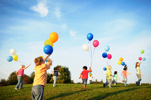Eight children in jeans and brightly colored shirts run across a grassy field on a sunny day.  Each of the children is holding two or three colorful balloons on strings.  The sky is clear but for a few small clouds.