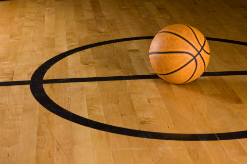 A basketball is reflected in the finish of the hardwood floor in a gym.
