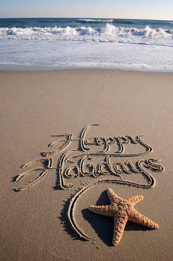 Happy Holidays message handwritten in the sand with a starfish and crashing waves in the background