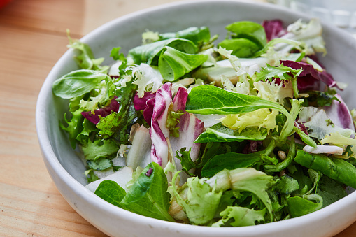 Delicious homemade gourmet healthy green salad bowl, with olive oil, garlic and goat cheese in olive oil, served in a rustic handmade ceramic bowl, with seasoning and herbs on a natural wooden kitchen or restaurant table, representing a healthy lifestyle, wellbeing and body care, side view with a copy space