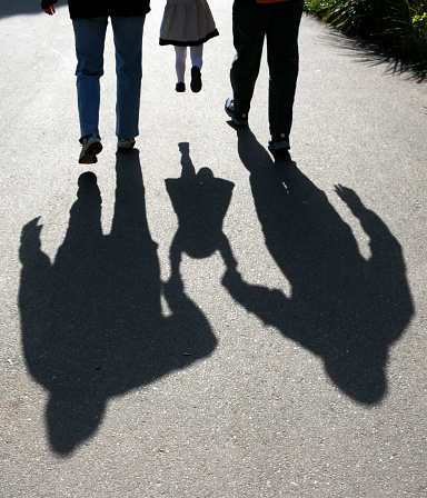 shadows behind two adults carrying a child on a sidewalk