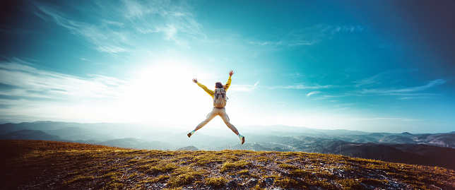 Happy woman with open arms jumping on mountain peak- Hiker with backpack celebrating success outdoor - People, success and sport concept
