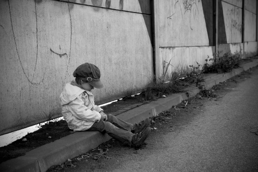 Depressed child sits on a side of a street,under concrete wall.