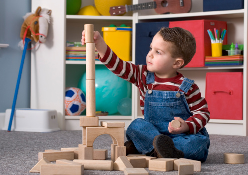 Shown here is a two-year-old boy happily playing with and looking at a tall stack of wooden toy blocks.  No visible logos. All toys are generic & non-branded.