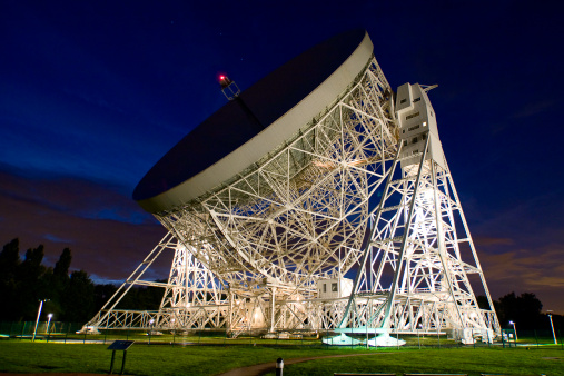 Jodrell Bank Observatory at night lit by floodlights with deep blue sky.