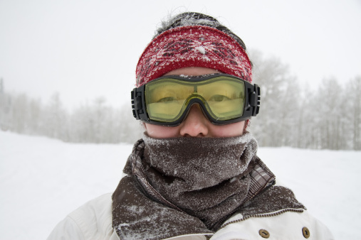 Girl or boy in ski helmet, sunscreen mask and balaclava close up stands against the backdrop of snow-covered mountain ski slope and a cloudy sky.
