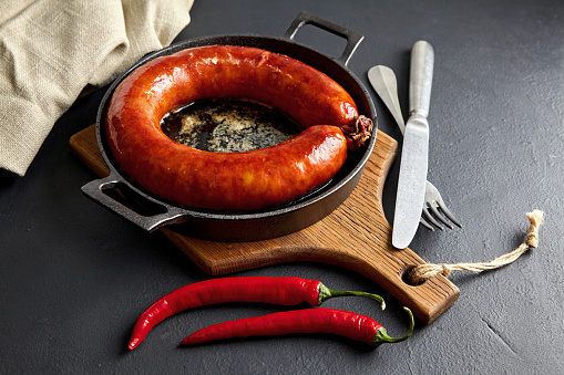 Fried sausage rolled in ring on cast-iron skillet and wooden cutting board. Grilled sausage with hot peppers and slice utensils.