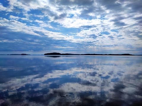 July morning cloudscape reflecting on the lake surface. White clouds and blue sky duplicating on almost mirror like water. Islands form a thin line in the horizon.