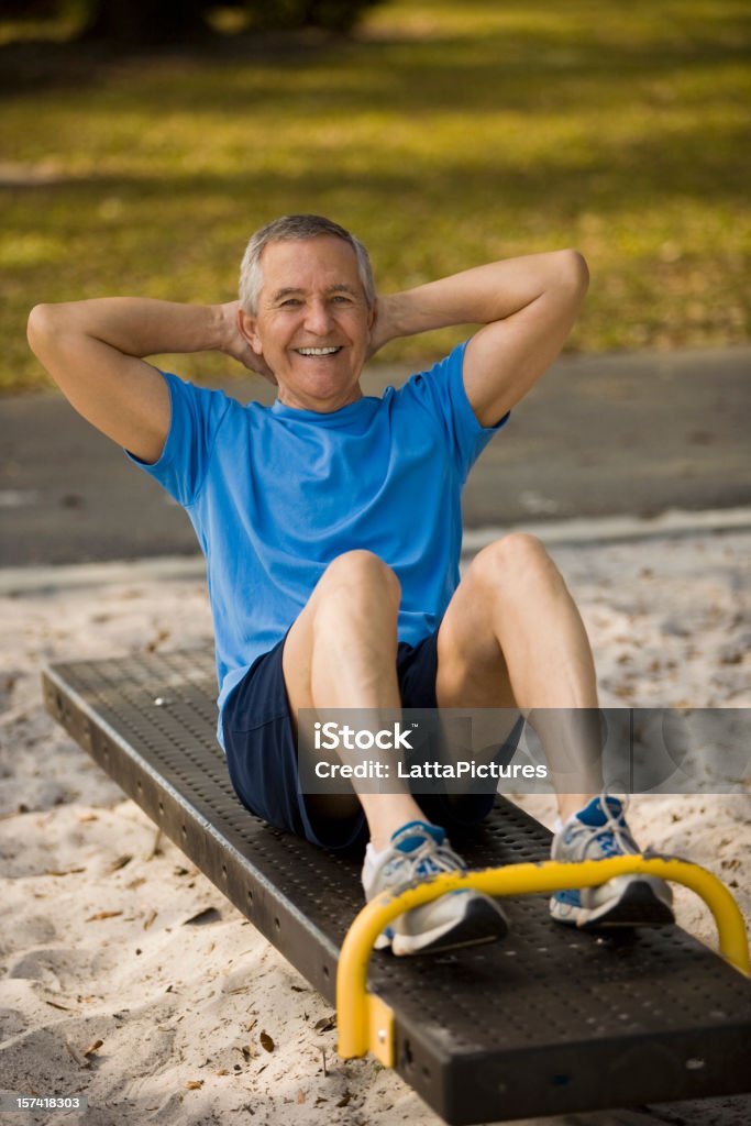 Uomo anziano seduto sulla panchina facendo Crunch "al park - Foto stock royalty-free di Abbigliamento sportivo