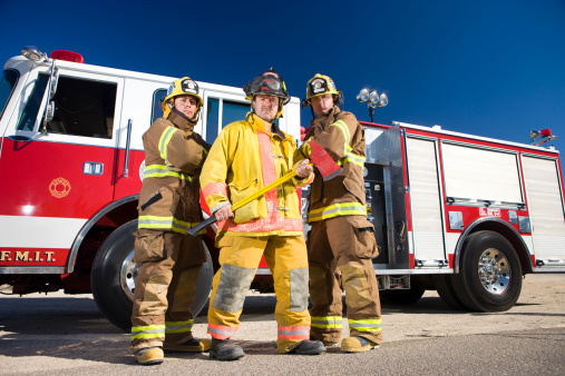 three real firefighters standing in front of a fire engine with authentic outfits/gear.