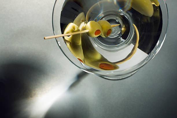 Martini Glass Cocktail Drink withToothpick Skewered  Olives Overhead View A chilled Martini garnished with two pimento stuffed green olives skewered by a toothpick, seen from a stylish overhead view. The hard liquor alcoholic beverage is a bar mixed drink and glamorous celebration party refreshment. Horizontal close-up view with copy space, on gray shadowy background with no people.  martini stock pictures, royalty-free photos & images