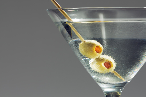 A chilled Martini garnished with two pimento stuffed green olives skewered by a toothpick, with condensation on the Martina glass surface. The hard liquor alcoholic beverage is a bar mixed drink and glamorous celebration party refreshment. Horizontal close-up view with copy space at left, on gray background with no people. 