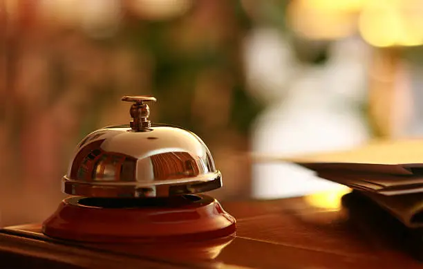 Close-up of a metal and red hotel service bell on a counter of a hotel reception. Business mail laying next to it. Light blur background of a lobby provides space for your text.  