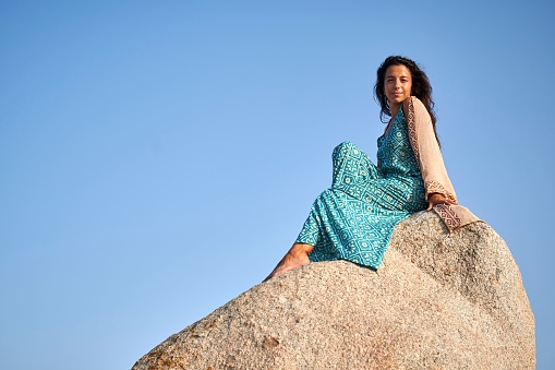 A young woman in a light-colored dress sitting atop a large rock formation, looking off into the distance