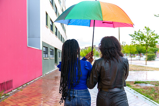 Two diverse friends under LGBT umbrella walk through the city in colorful rain, embracing their differences.